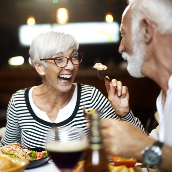 An older woman with gray hair and glasses laughs while dining with a gray-haired man. They are sharing food at a restaurant, and drinks and plates of food are visible on the table.