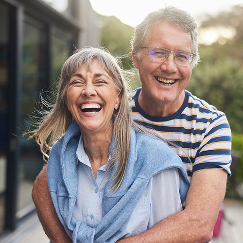 An older couple smiles and laughs outdoors. The woman is in front, wearing a blue shirt and a sweater over her shoulders, and the man stands behind her with his arms around her, wearing glasses and a striped shirt.