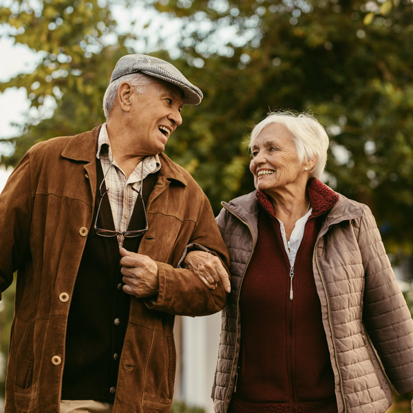 An elderly man and woman walk arm in arm outdoors, smiling at each other. They are dressed in warm jackets and appear happy, with green trees in the background.