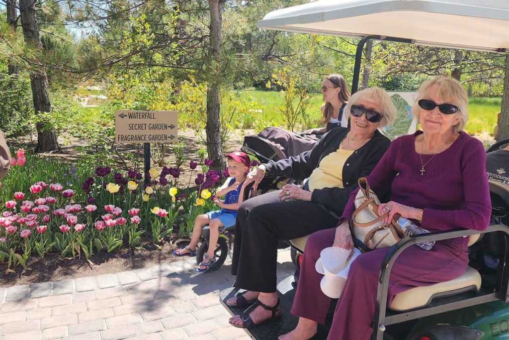Two older women sit on a golf cart beside a young child in a stroller near blooming tulips. A sign points to a waterfall, secret garden, and fragrance garden. Trees and greenery fill the background.