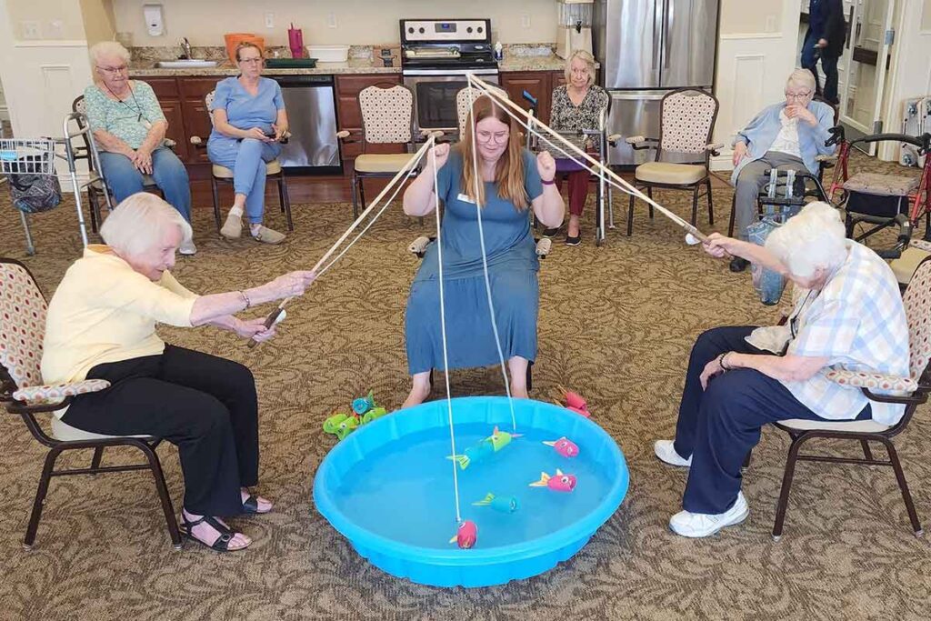 A group of elderly women and a younger woman sit in a circle, playing a fishing game with toy rods and colorful plastic fish in a small blue kiddie pool indoors.