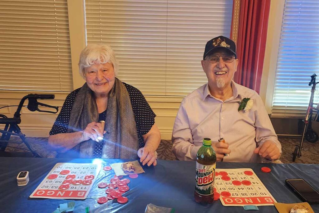 Two smiling elderly people sit at a table playing bingo. The woman holds a flashlight and bingo card, while the man wears a hat and has a bottle of Mountain Dew in front of him. Bingo chips and cards are spread on the table.