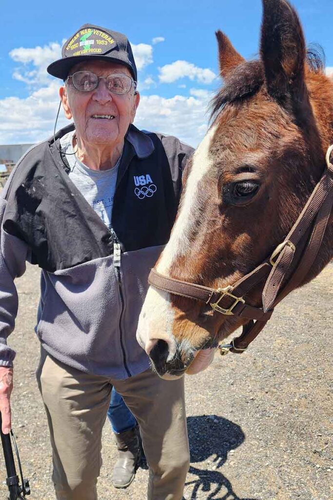 An elderly man wearing glasses, a black USA Olympic jacket, and a cap that says "World War II Veteran" stands outdoors smiling next to a brown and white horse under a blue sky with scattered clouds.