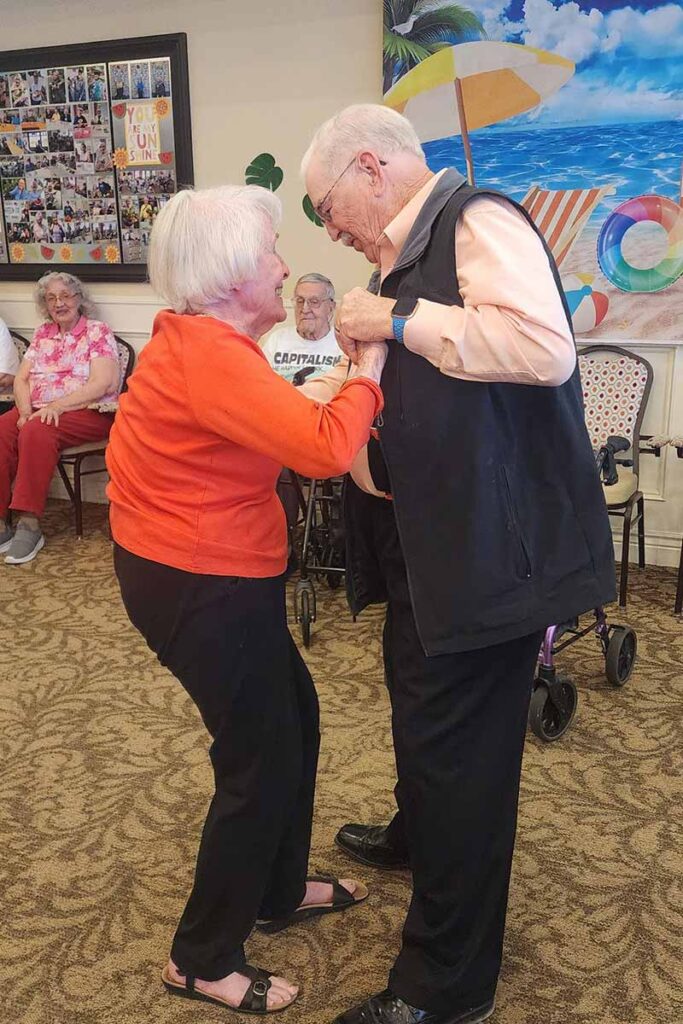 An elderly man and woman smile and hold hands while dancing together in a bright, cheerful room with colorful beach-themed decorations and other seniors seated in the background.