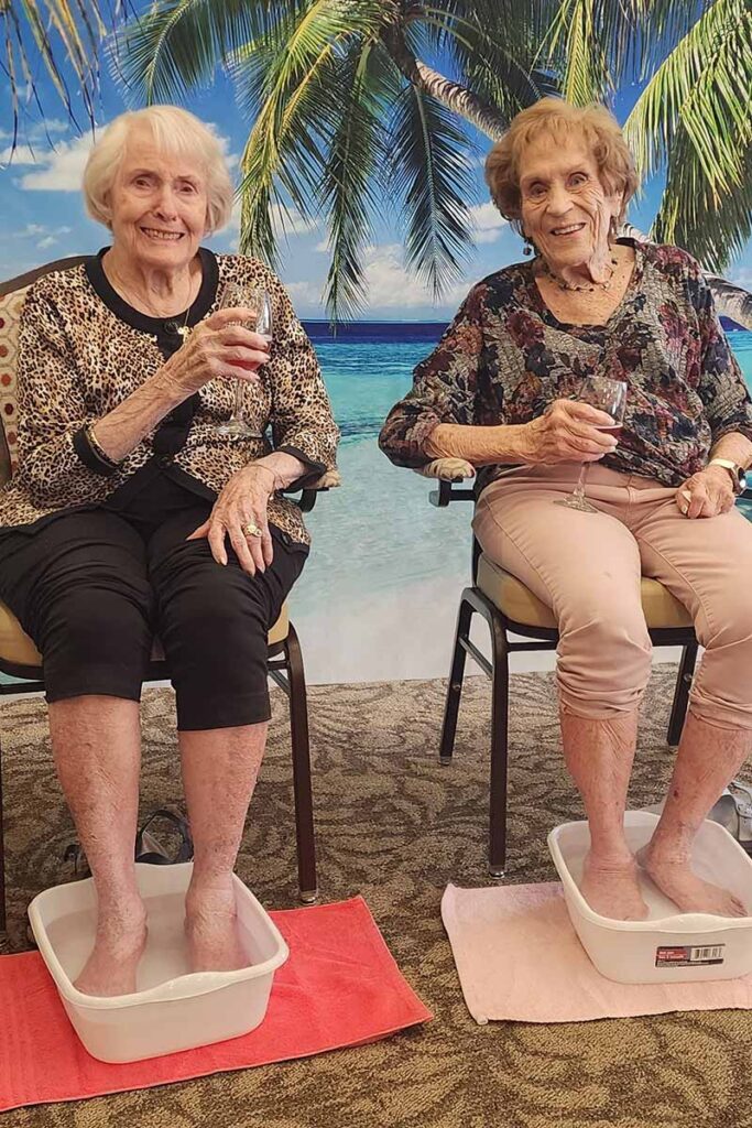Two elderly women sit on chairs with their feet soaking in tubs of water, each holding a glass of wine. They are smiling, with a tropical beach backdrop and palm trees behind them.