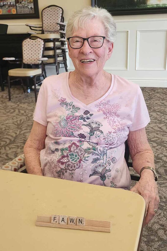 An elderly woman with short white hair and glasses smiles while sitting at a table. In front of her is a wooden tile holder with Scrabble tiles spelling "FAWN." She wears a light pink, floral-patterned shirt.