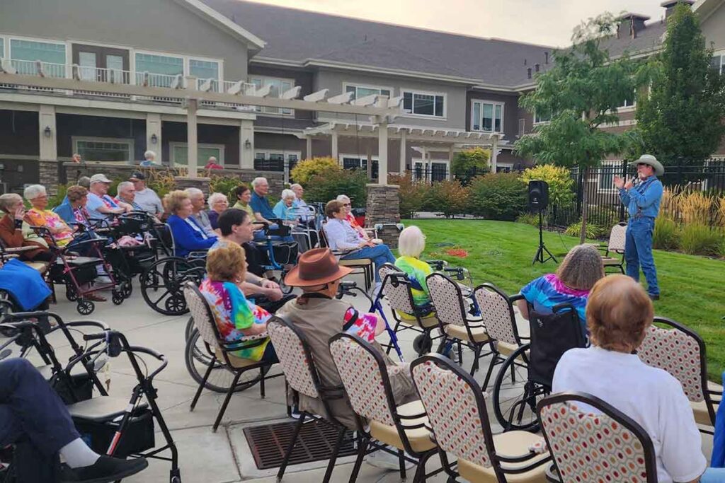 A group of elderly people, some in wheelchairs, sits outdoors facing a man in denim and a cowboy hat who is performing or speaking. The event takes place at a care facility with garden and patio seating.