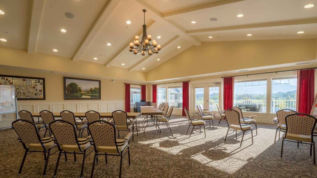 A spacious, sunlit meeting room with patterned chairs arranged in a semi-circle, large windows with red curtains, a chandelier, and a landscape painting on the wall.