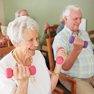 Two smiling older adults exercise indoors, sitting on chairs and lifting small dumbbells. The woman holds pink weights and wears a white blouse, while the man holds purple weights and wears a plaid shirt. Others are visible in the background.