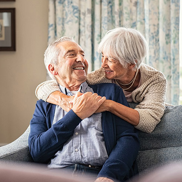 An elderly couple sits on a couch at home, smiling and embracing lovingly. The woman hugs the man from behind, and both appear happy and relaxed in a cozy, well-lit room with patterned curtains.