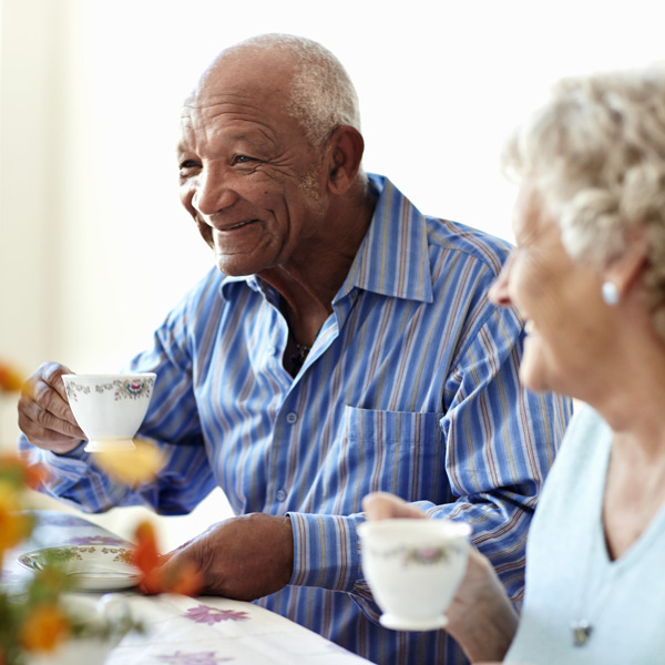 Two older adults sit together at a table, smiling and holding teacups. The man wears a blue striped shirt, and the woman wears a light blue top. The atmosphere appears warm and friendly.