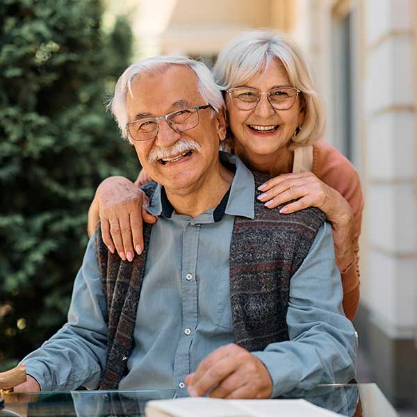 An elderly couple smiles warmly outdoors. The man sits at a table with a book, wearing glasses and a gray shirt, while the woman stands behind him, resting her hands on his shoulders, also smiling and wearing glasses.