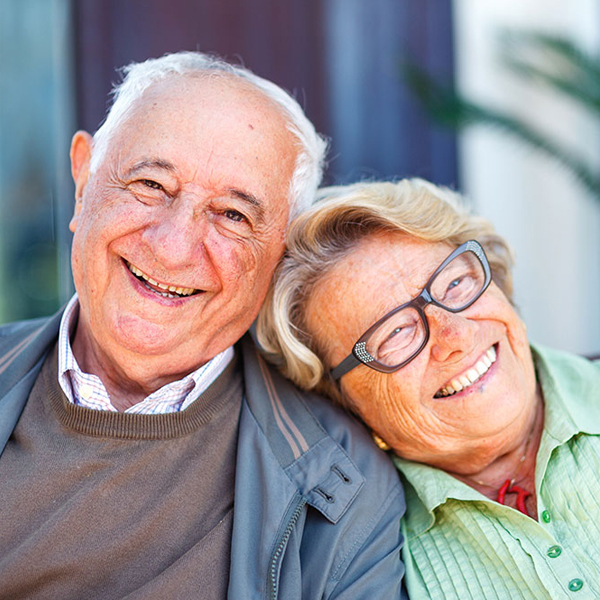 An elderly man and woman smiling warmly, with the woman resting her head on the man's shoulder. Both appear happy and relaxed, and are dressed in casual clothing. The background is softly blurred.