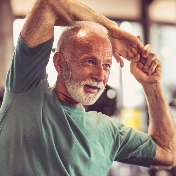 An older man with gray hair and a beard, wearing a green t-shirt, is stretching his arms above his head and smiling in a gym or fitness center.