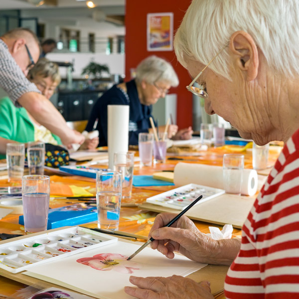 A group of elderly people sits around a table, painting with watercolors. Art supplies and glasses of water are scattered on the table, and one woman in the foreground paints a flower on paper.