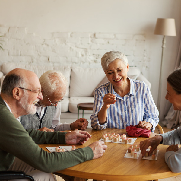 Four older adults sit around a wooden table, smiling and laughing while playing a board game together in a cozy, well-lit living room.