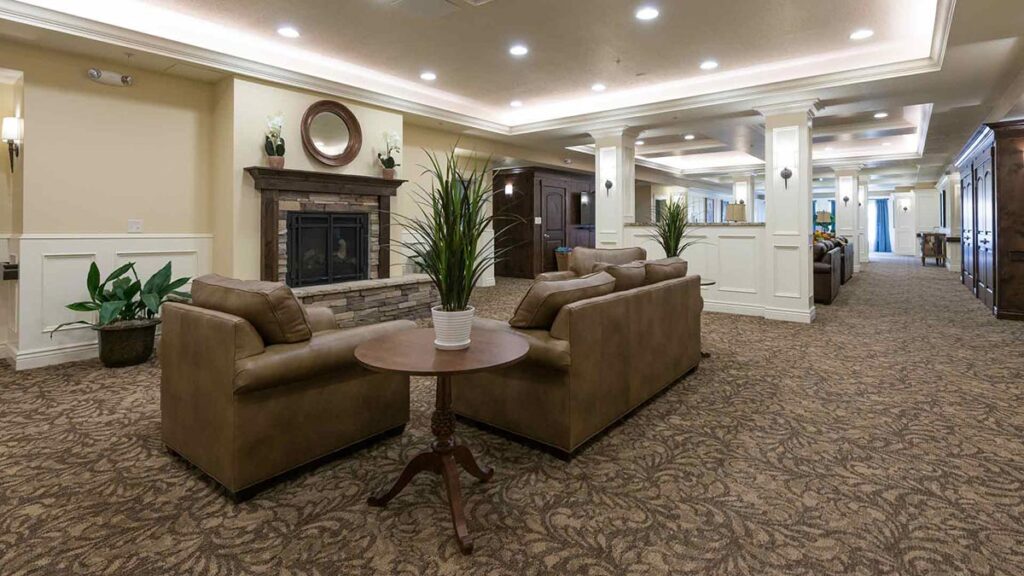 A spacious, well-lit lounge with brown sofas, a round wooden table, decorative plants, carpeted floor, and a stone fireplace along the wall, giving the room a warm, inviting atmosphere.