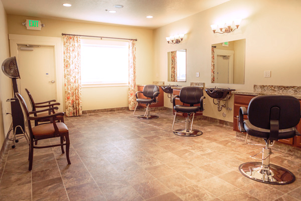 A bright, tidy salon room with three black salon chairs facing mirrors, two brown waiting chairs, a hair dryer station, tiled floor, floral curtains on a window, and warm lighting.