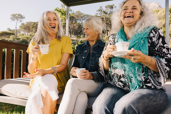 Three older women with gray hair sit closely together on an outdoor bench, laughing and holding teacups. They appear joyful, enjoying each other's company on a sunny day with greenery in the background.