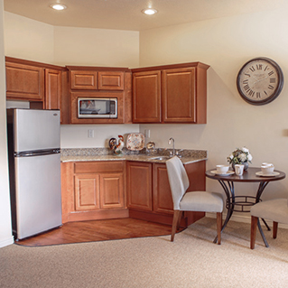 A small kitchen with wooden cabinets, a stainless-steel fridge, a microwave, and granite countertops. A round table with two chairs is set with cups and a flower arrangement. A large wall clock hangs nearby.