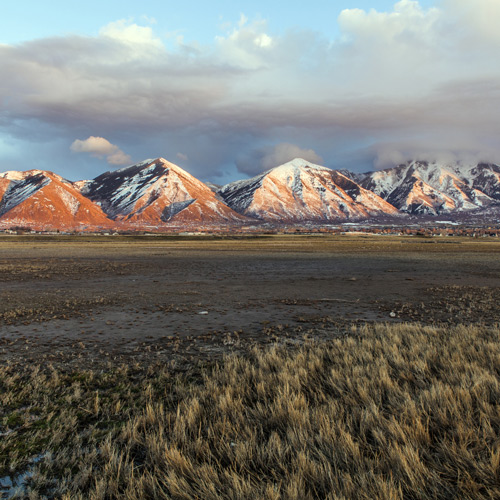 Snow-capped mountains glow with orange light at sunset, rising behind a flat, grassy plain under a partly cloudy sky.