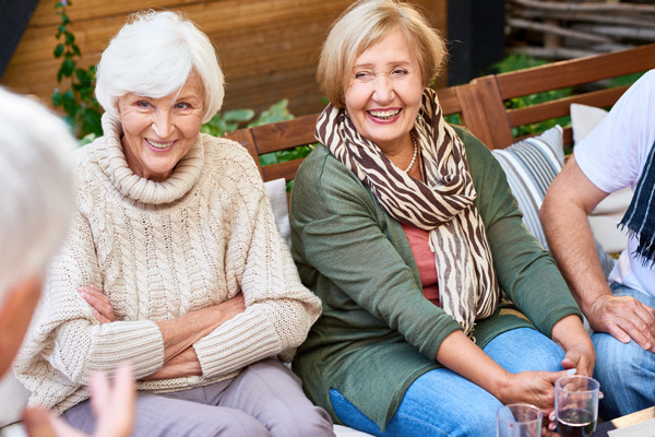 Two older women sit together on a bench outdoors, smiling and talking. One wears a beige knit sweater, the other a green top with a striped scarf. They are joined by others whose faces are partly out of frame.
