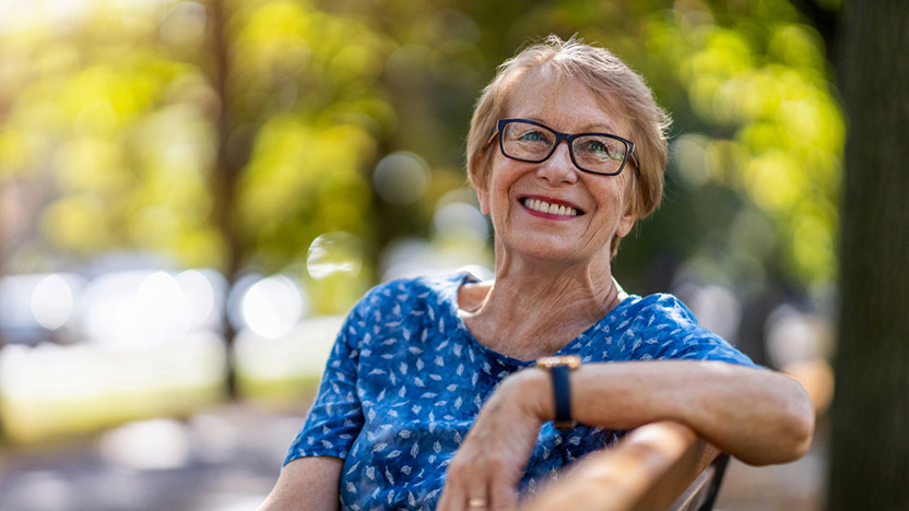 Smiling older woman wearing glasses and a blue patterned shirt sits on a park bench, enjoying a sunny day with green trees—embracing her transition to assisted living amid soft sunlight in the background.