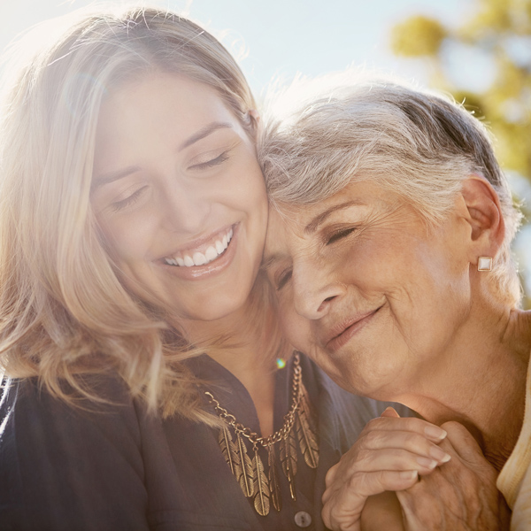 A smiling young woman and an older woman with gray hair share a close, affectionate embrace outdoors in sunlight, both appearing happy and content.