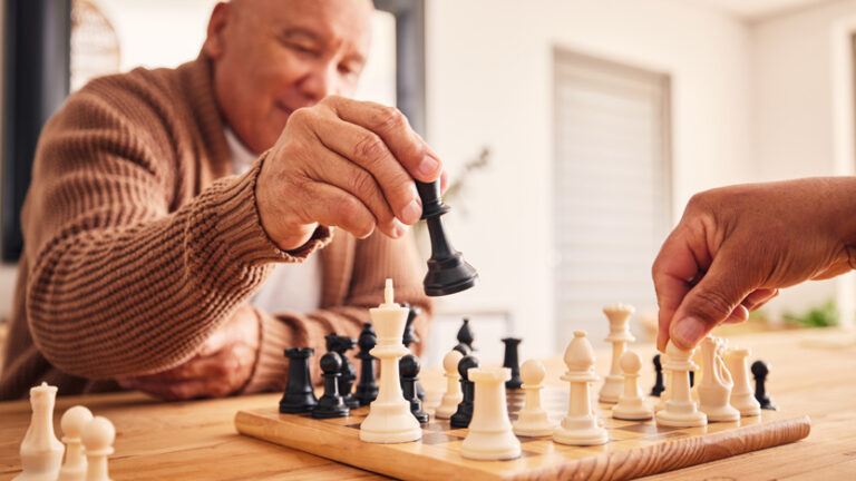 An older adult wearing a brown sweater is moving a black chess piece during a chess game, enjoying meaningful moments as they transition to assisted living, while another person's hand prepares to move a white piece on the wooden chessboard.