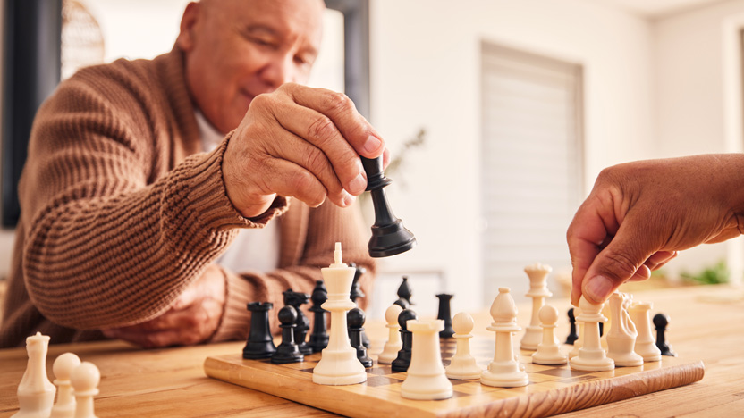 An older adult wearing a brown sweater is moving a black chess piece during a chess game, enjoying meaningful moments as they transition to assisted living, while another person's hand prepares to move a white piece on the wooden chessboard.